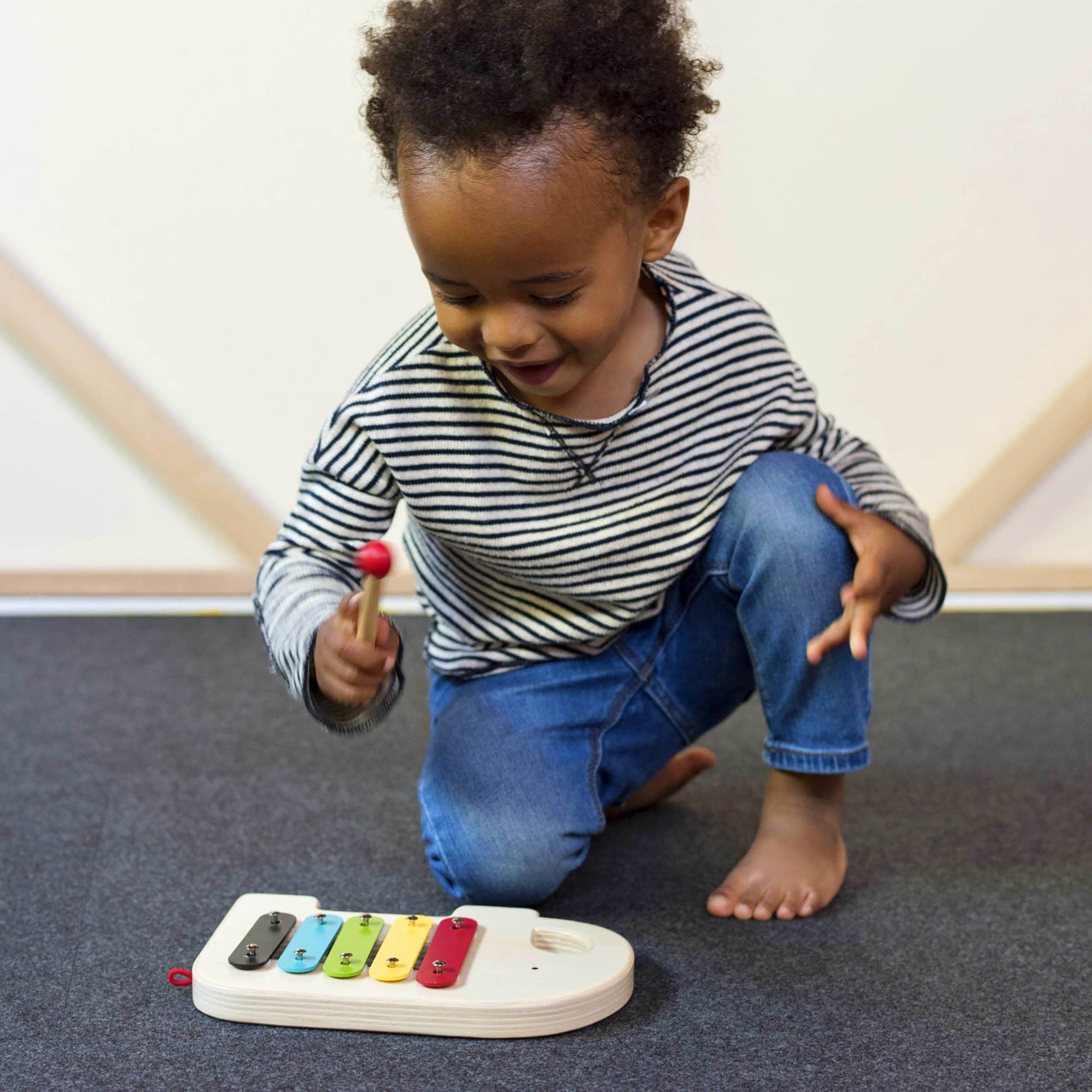 A young child in a striped shirt and jeans plays with the Petit Collage Wooden Elephant Xylophone on a carpeted floor, holding a red mallet.