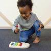 A young child in a striped shirt and jeans plays with the Petit Collage Wooden Elephant Xylophone on a carpeted floor, holding a red mallet.