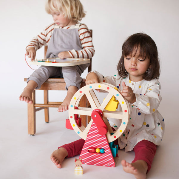 Two young children play; one sits on the floor with the Petit Collage Wooden Ferris Wheel Carnival Play Set, while the other sits in a chair with a bead maze.