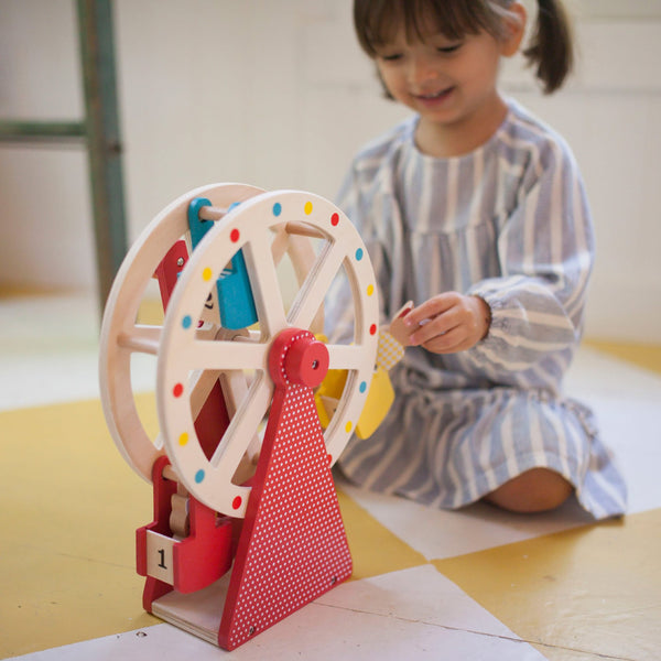 A young child in a striped dress sits on the floor, playing with the Petit Collage Wooden Ferris Wheel Carnival Play Set.