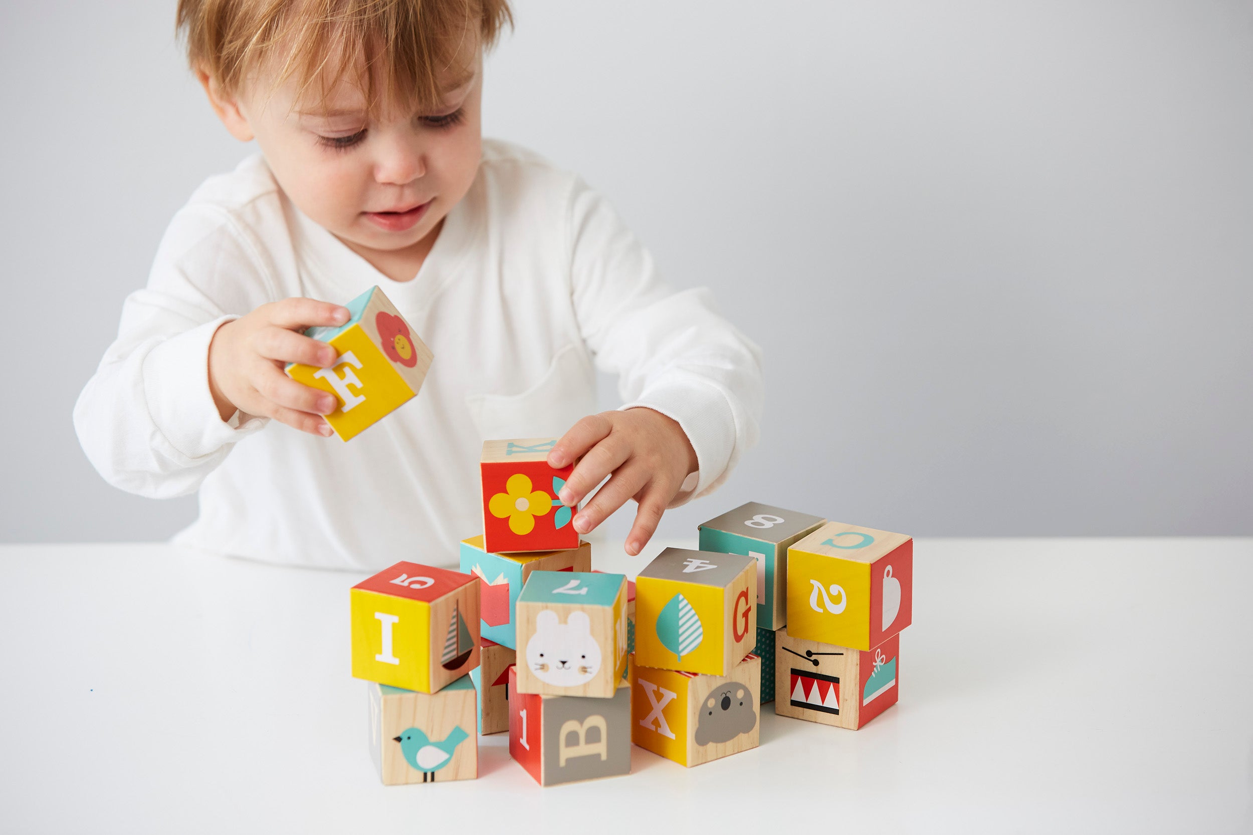 A young child builds with Petit Collage ABC Wooden Alphabet Blocks, stacking colorful wooden letters and animal blocks on a white table.