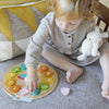 A young child sits on a couch holding a stuffed toy and playing with the Petit Collage Multi-Language + Counting + Colors Wooden Learning Clock, featuring colorful numbered pieces.