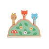 The Petit Collage Petit Friends Wooden Pop-Up Toy features cat, fox, and bear figures popping from a green base adorned with leaves, flowers, and a red mushroom.