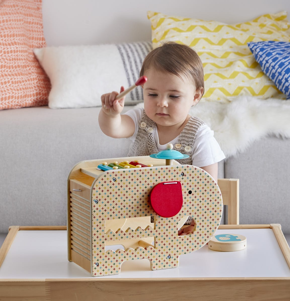 A toddler plays with the Petit Collage Wooden 5-in-1 Elephant Music Toy, striking colorful keys with a mallet while seated at a table in the living room.