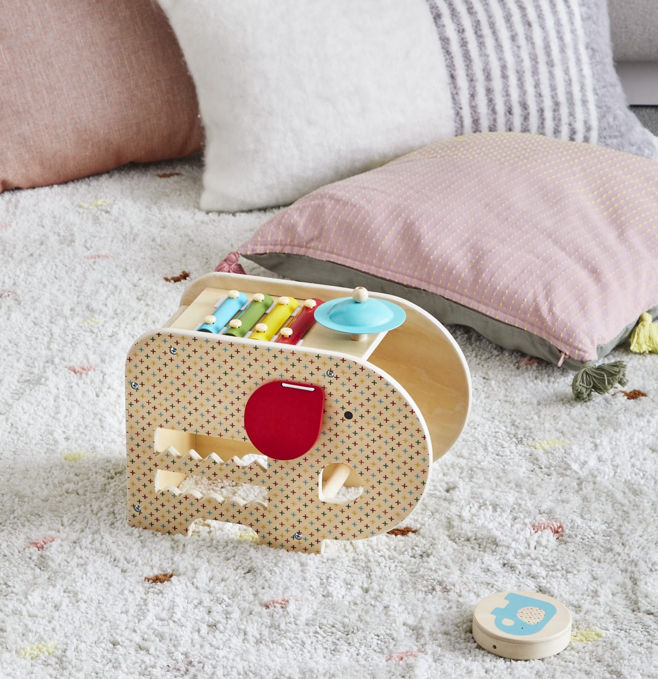 The Petit Collage Wooden 5-in-1 Elephant Music Toy, featuring a xylophone, cymbal, and maze, rests on a plush carpet beside two pastel-colored pillows.