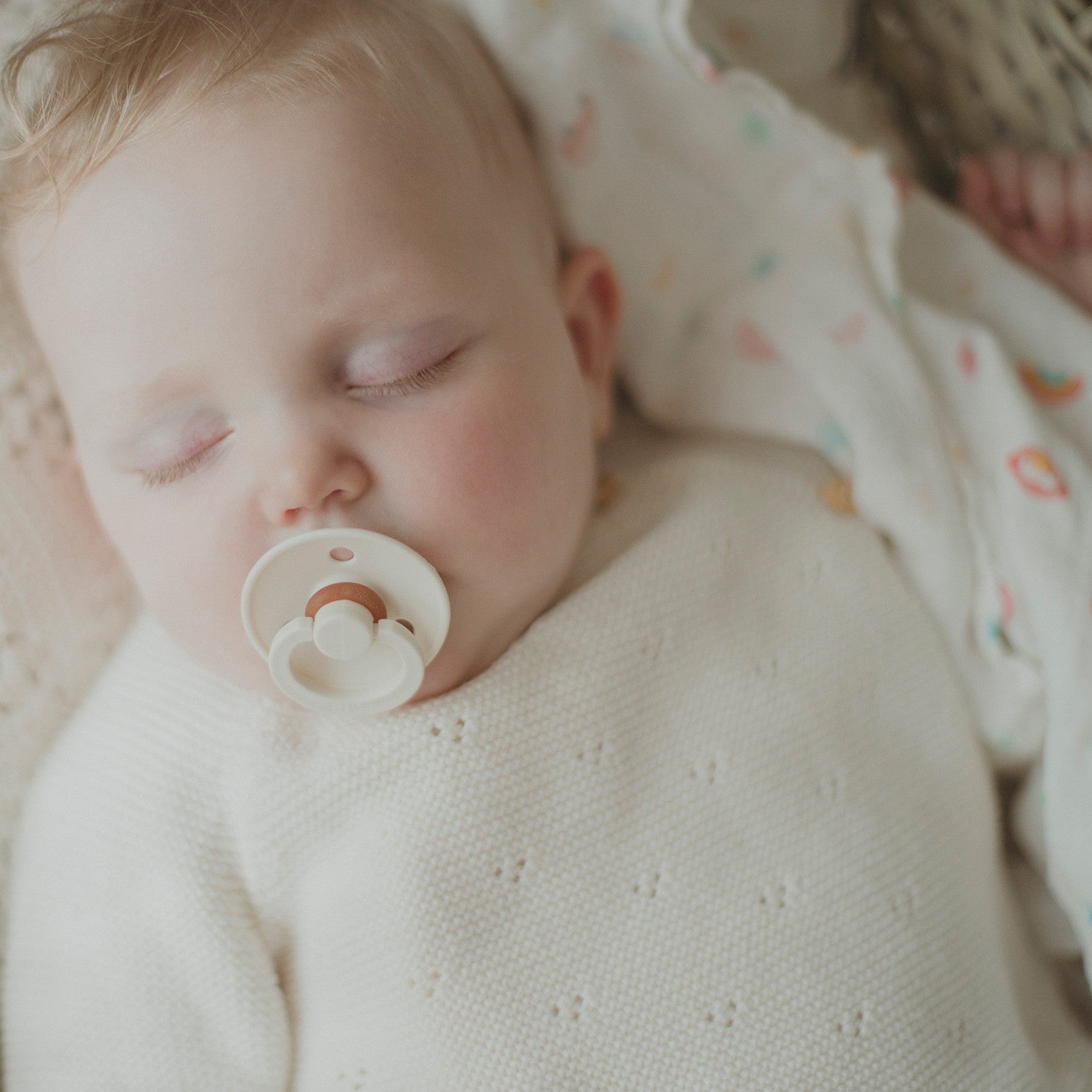 A baby with a pacifier sleeps peacefully, dressed in the Petit Collage Little Monkey Baby Gift Set and lying on a patterned blanket.