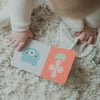 A baby in white sits on a rug, holding an open board book from the Petit Collage Little Monkey Baby Gift Set, featuring simple illustrations of a bear and a flower.