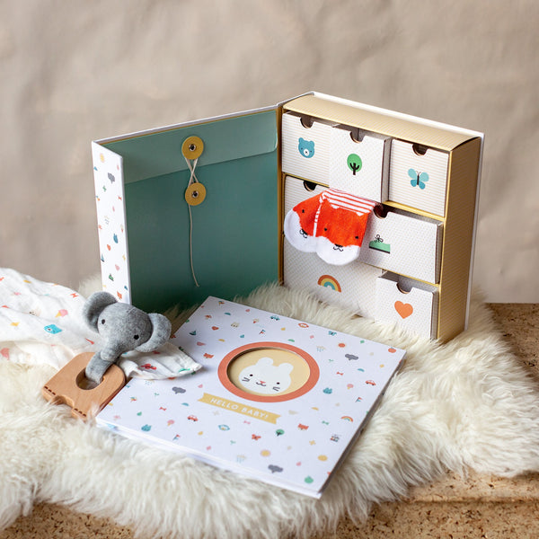 The Petit Collage Baby Memory Box is a gift set featuring small keepsake drawers, a plush elephant teether, muslin cloth, and a Hello Baby scrapbook, all displayed on a soft surface.