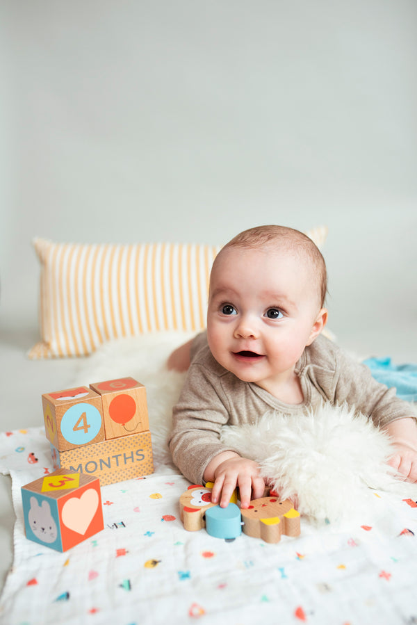 A baby rests beside Petit Collage Wooden Baby Milestone Blocks showing 4 months, with small toys and a striped pillow in the background.
