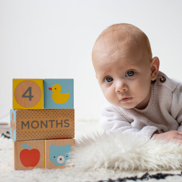 A baby rests on a white furry rug next to Petit Collage Wooden Baby Milestone Blocks showing 4 months with images of a duck, apple, and bear.