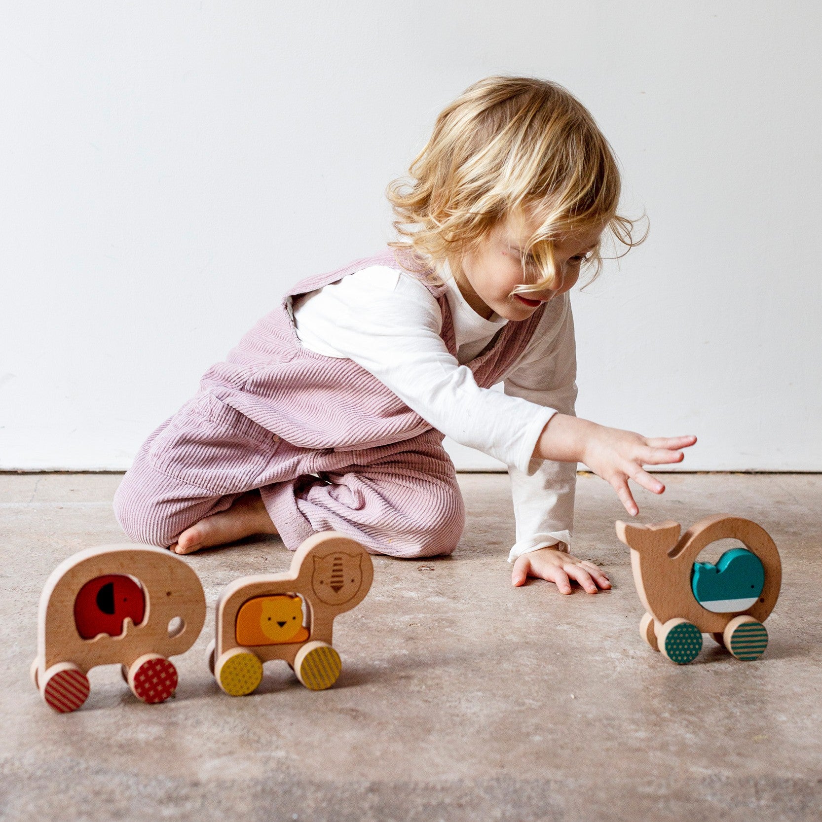 A young child in pink overalls plays on the floor with the Petit Collage Wooden Push Along Whale Toy and two other animal-shaped wooden toys—a lion and an elephant.