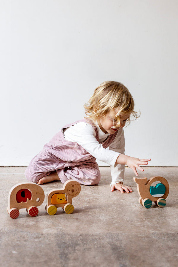 A young child in pink overalls plays on the floor with the Wooden Push Along Lion Toy from Petit Collage.