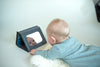 A baby in a light blue outfit lies on a soft white rug, gazing at their reflection in the Petit Collage Tummy Time Peekaboo mirror propped up close by.