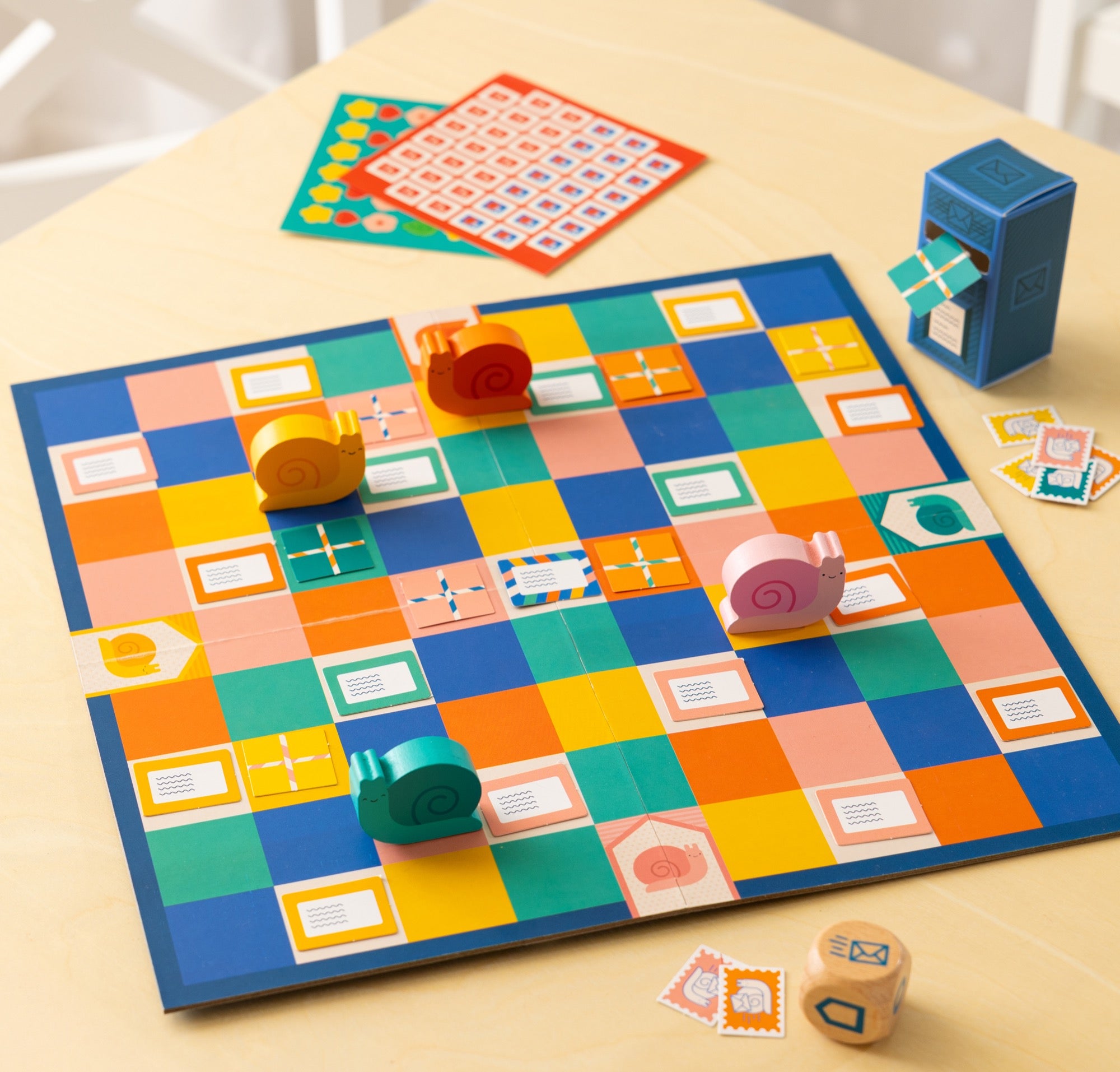 Snail Express by Petit Collage is a colorful board game featuring a checkered board, four snail-shaped pieces, dice, cards, and a blue mailbox figure, all displayed on a light wooden table.