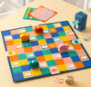 Snail Express by Petit Collage is a colorful board game featuring a checkered board, four snail-shaped pieces, dice, cards, and a blue mailbox figure, all displayed on a light wooden table.