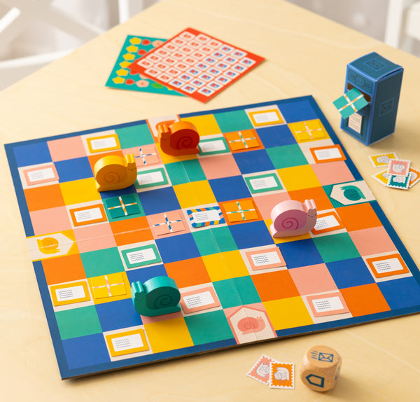 Snail Express by Petit Collage is a colorful board game featuring a checkered board, four snail-shaped pieces, dice, cards, and a blue mailbox figure, all displayed on a light wooden table.