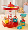 The Petit Collage Merry-Go-Round Wooden Carousel showcases animal figures and numbered blocks, surrounded by extra colorful blocks. In the blurred background, a teddy bear and a blue toy box can be seen.