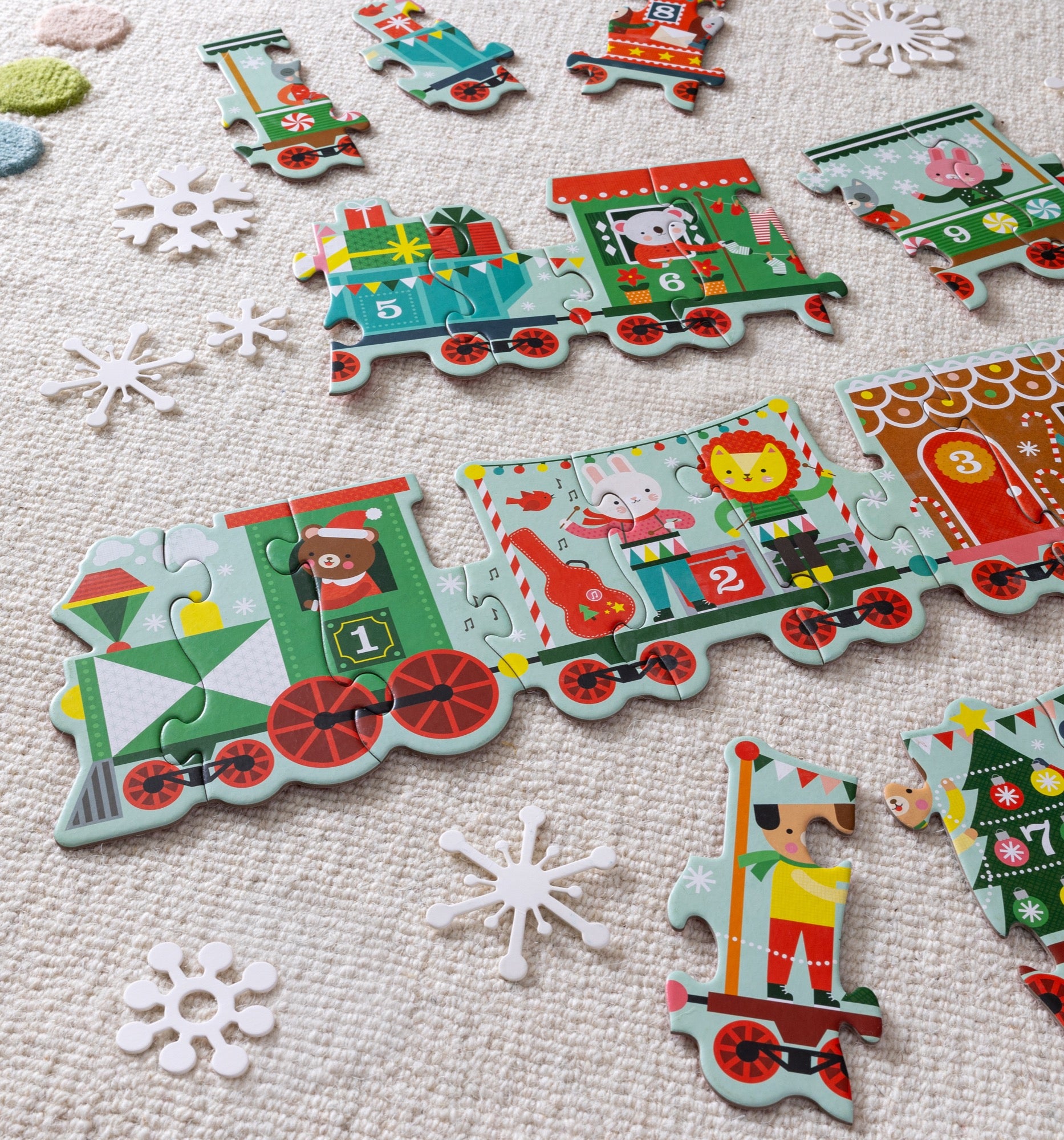 The Petit Collage Christmas Train Shaped Floor Puzzle, featuring vibrant animal designs, is arranged on a textured rug with loose pieces and scattered white paper snowflakes nearby.