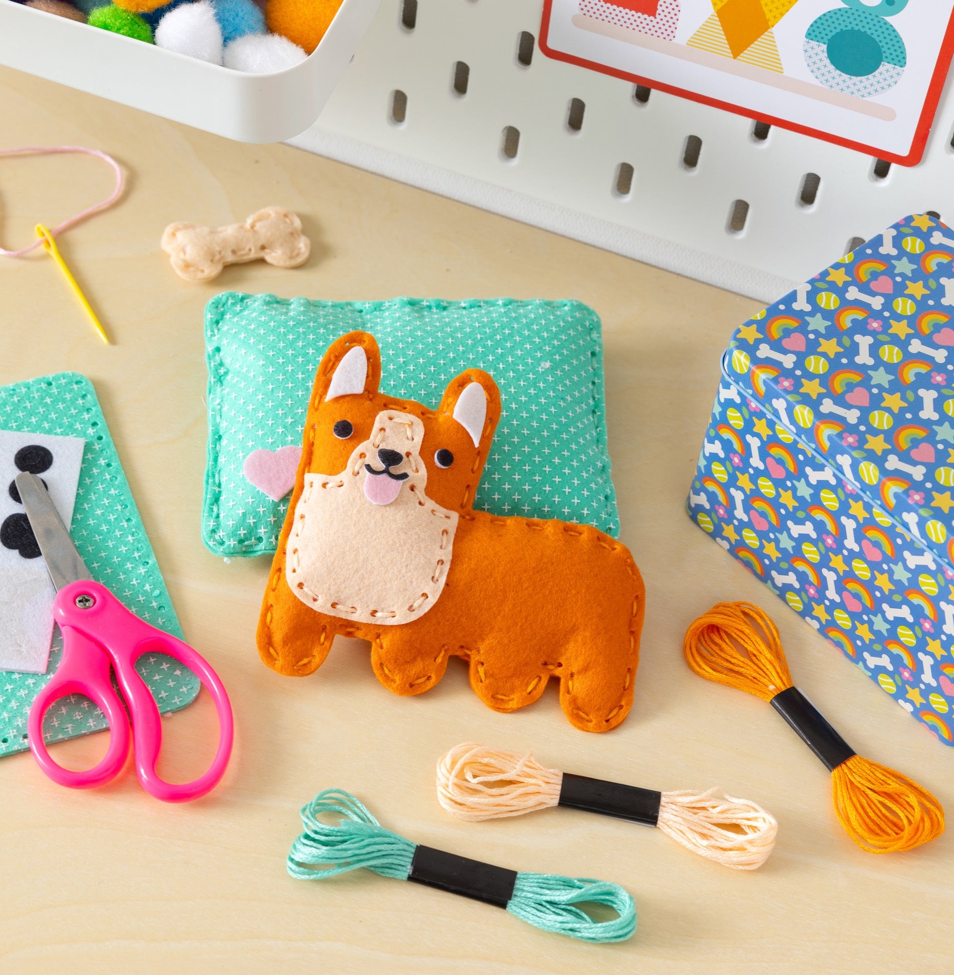 A Mini Craft Kit: Pocket Pet Corgi by Petit Collage rests on a teal pillow with embroidery floss, scissors, a needle, and a blue patterned box nearby. Craft supplies and a pegboard appear in the background.