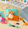 A Mini Craft Kit: Pocket Pet Corgi by Petit Collage rests on a teal pillow with embroidery floss, scissors, a needle, and a blue patterned box nearby. Craft supplies and a pegboard appear in the background.