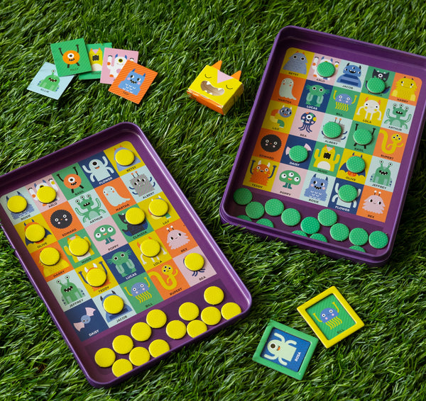 The Petit Collage Whos That Monster? Magnetic Travel Game features two colorful monster-themed bingo boards, yellow and green markers, matching monster cards, and a small yellow box with a cartoon face.
