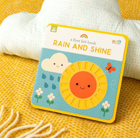 A First Felt Book: Rain and Shine by Petit Collage rests on a yellow textured blanket, with a white and blue cushion in the background.