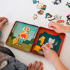 Two children arrange Petit Collage Animal Band On-The-Go Magnetic Play Set magnets of animals and instruments on illustrated playsets inside an open tin, with extra magnets scattered on the table.
