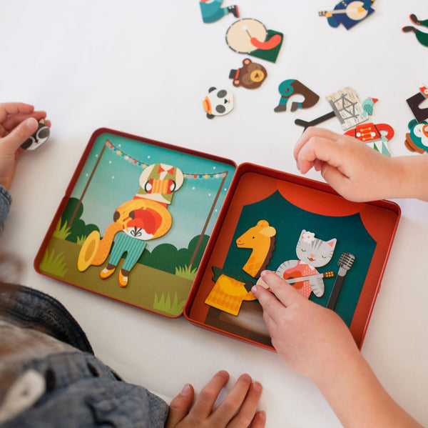 Two children arrange Petit Collage Animal Band On-The-Go Magnetic Play Set magnets of animals and instruments on illustrated playsets inside an open tin, with extra magnets scattered on the table.