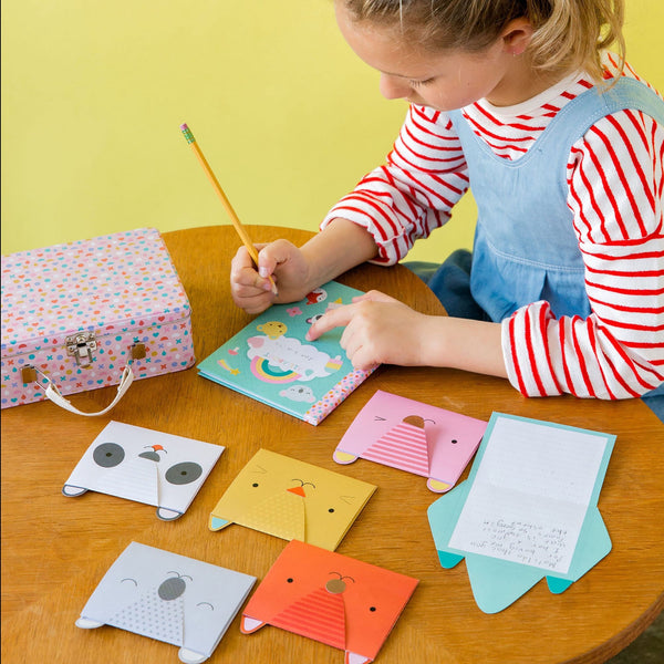 A child writes in a notebook at a table with Petit Collage’s Animal Pals My Stationery Set, featuring colorful animal-shaped cards, a polka dot box, and a closed pencil.