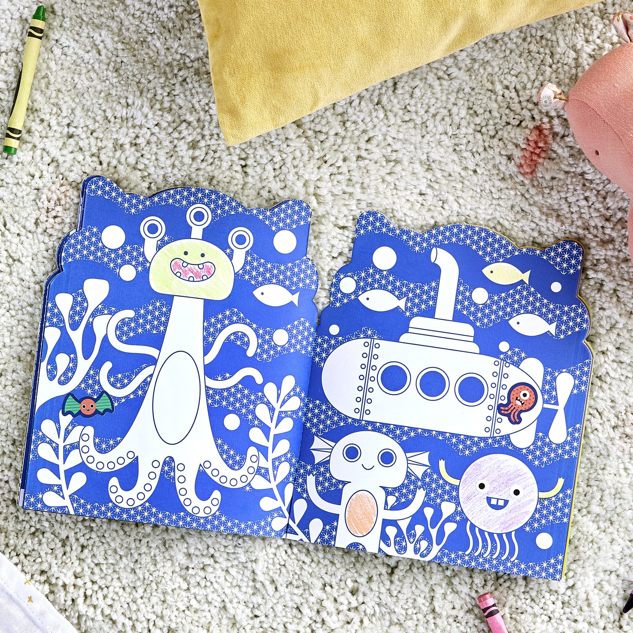 The Coloring Book with Stickers: Monsters by Petit Collage lies open, showing playful monster illustrations. Crayons and toys are scattered on the carpet around the book.