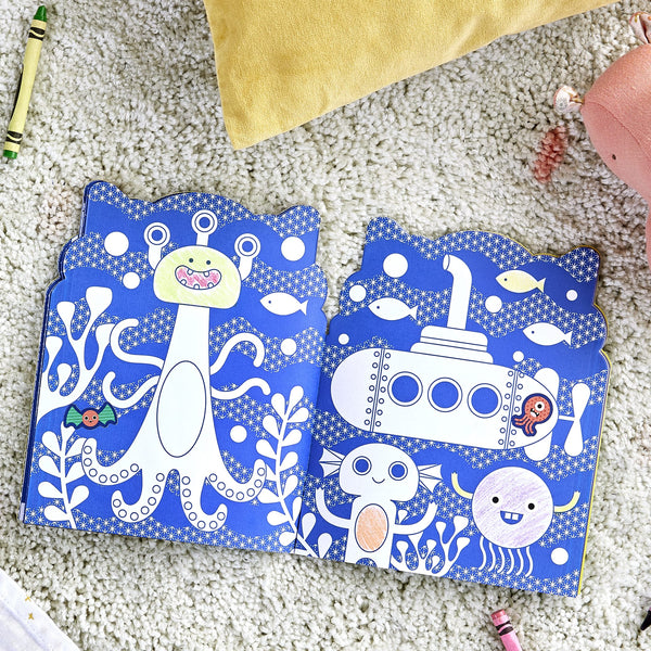 The Coloring Book with Stickers: Monsters by Petit Collage lies open, showing playful monster illustrations. Crayons and toys are scattered on the carpet around the book.