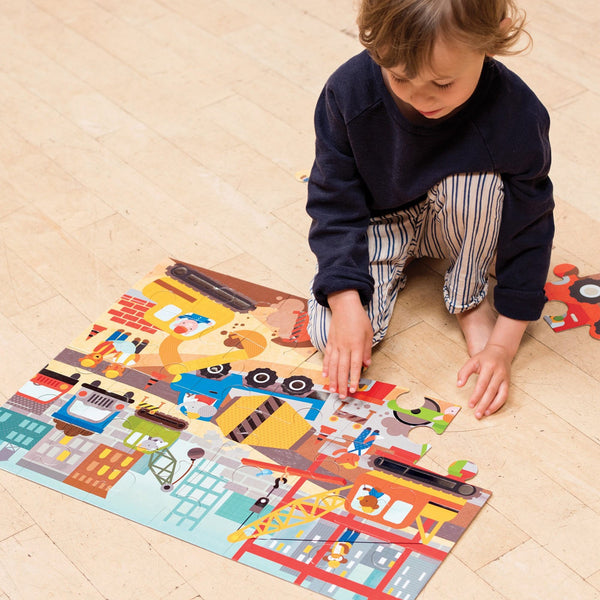 A young child assembles the Petit Collage Construction Site 24-Piece Floor Puzzle on a wooden floor.