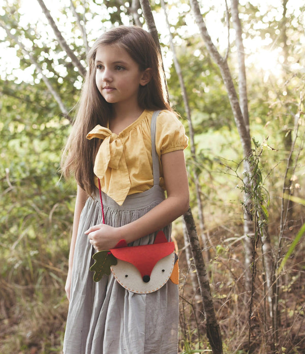 A young girl with long brown hair stands in a wooded area, wearing a yellow blouse, gray skirt, and carrying the Petit Collage DIY Woodland Fox Felt Bag.
