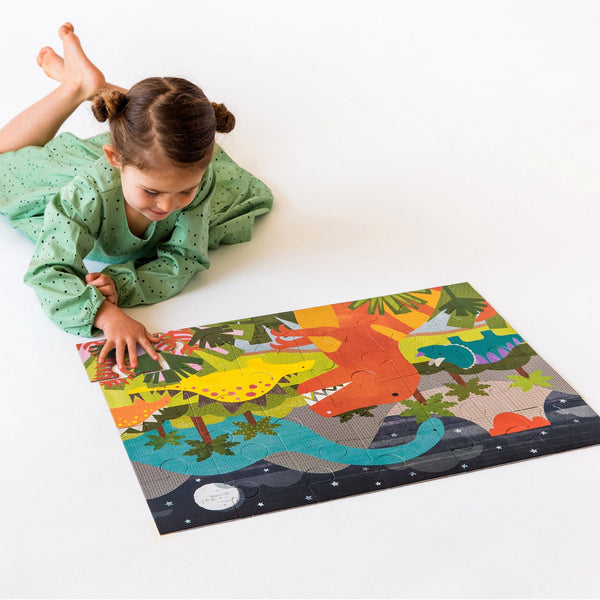 A young girl in a green dress assembles the colorful Petit Collage Dinosaur Kingdom 24-Piece Floor Puzzle on the floor.