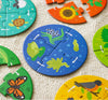 The Petit Collage Discovery Puzzle: Life Cycle features circular animal-themed pieces with illustrations of a frog, fish, turtle, butterfly, bird, and plants, shown assembled and scattered on a textured surface.