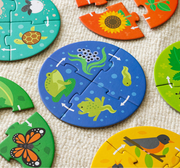The Petit Collage Discovery Puzzle: Life Cycle features circular animal-themed pieces with illustrations of a frog, fish, turtle, butterfly, bird, and plants, shown assembled and scattered on a textured surface.