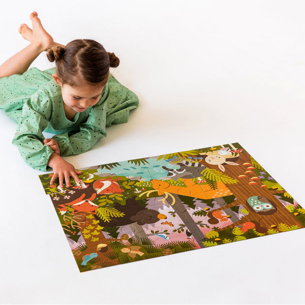 A young girl in a green dress lies on the floor, assembling the Petit Collage Enchanted Woodland 24-Piece Floor Puzzle, a colorful animal-themed jigsaw, on a white surface.