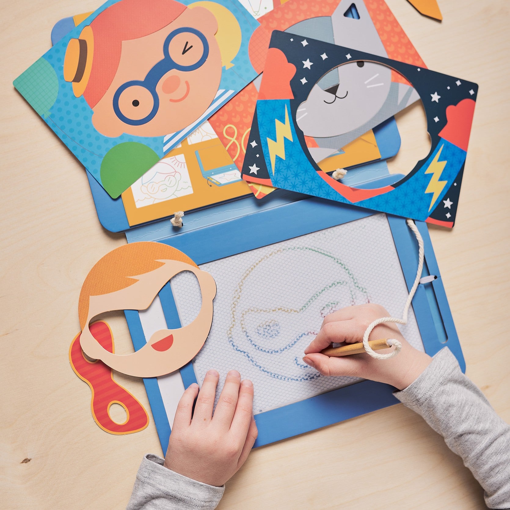 A child traces a superhero face on the Petit Collage Funny Faces Magic Sketch Board, surrounded by colorful character templates and masks on a light wooden table.