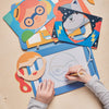 A child traces a superhero face on the Petit Collage Funny Faces Magic Sketch Board, surrounded by colorful character templates and masks on a light wooden table.