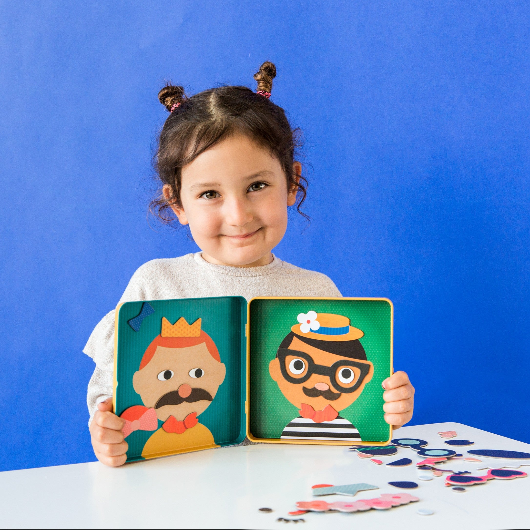 A young child with dark hair in buns holds up an open Petit Collage Funny Faces Magnetic Play Set, showing two illustrated faces and various magnetic pieces on the table.