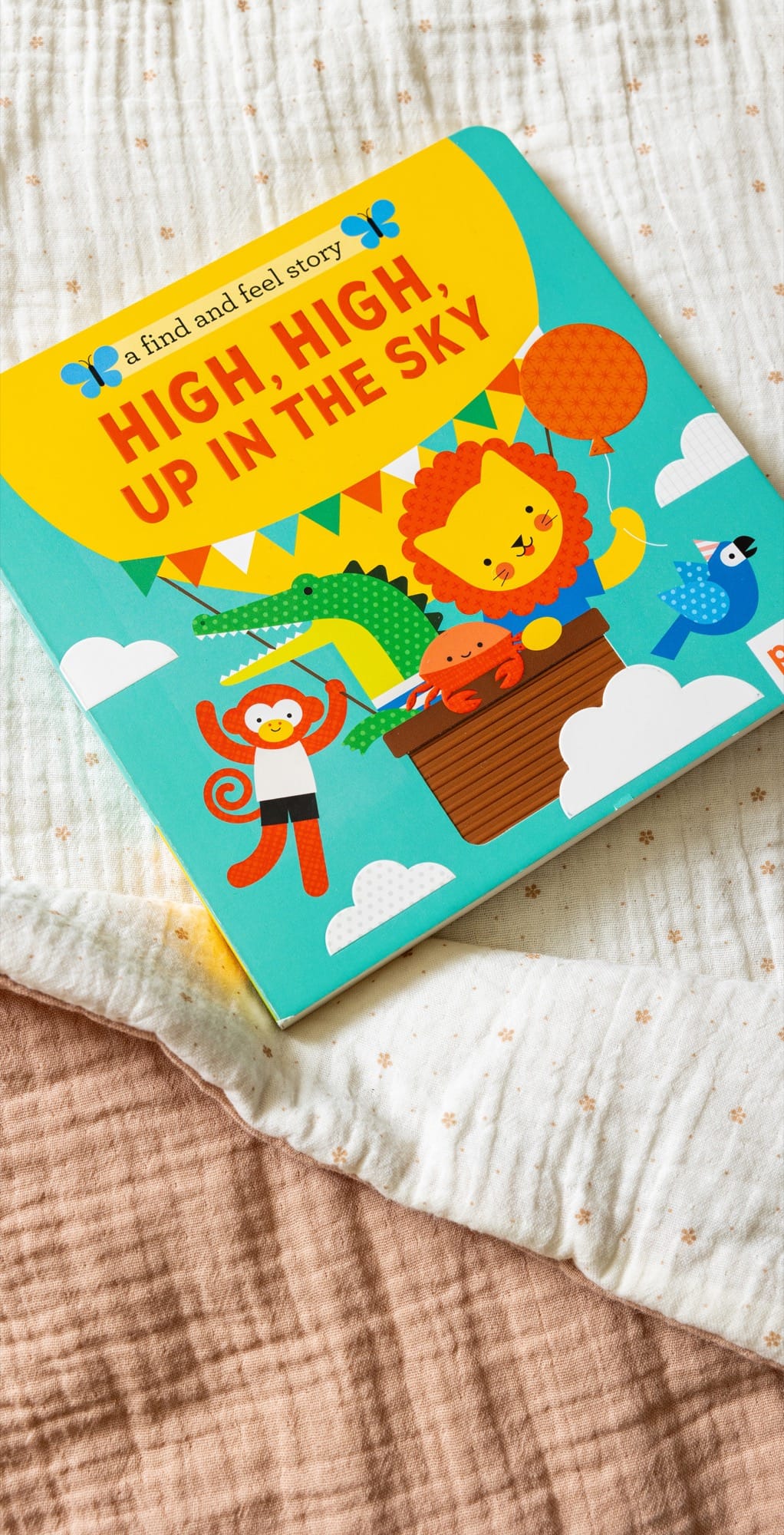 The Petit Collage book A Find and Feel Story: High, High, Up in the Sky features cartoon animals in a hot air balloon, shown on a textured fabric surface.