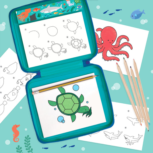 The Petit Collage "How to Draw Ocean Life" kit features step-by-step guides for sketching sea animals like a turtle, octopus, shark, and whale with colored pencils on an ocean-themed blue background.
