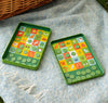 Two Petit Collage In-the-Park Bingo Magnetic Travel Game boards, featuring colorful tiles and round playing pieces, are set out on a blue textured picnic blanket.