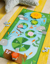The Petit Collage Leaping Frogs! children’s board game, featuring animal pieces and tokens, is set up on a yellow-striped surface next to a textured star-shaped pillow.