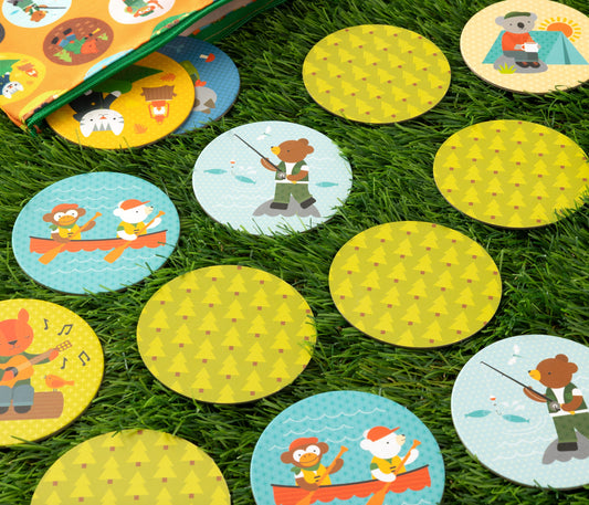 The Little Campers Matching Game by Petit Collage features round cards with illustrated animals camping, fishing, playing music, and boating, along with cards showing repeating tree patterns spread on green grass.
