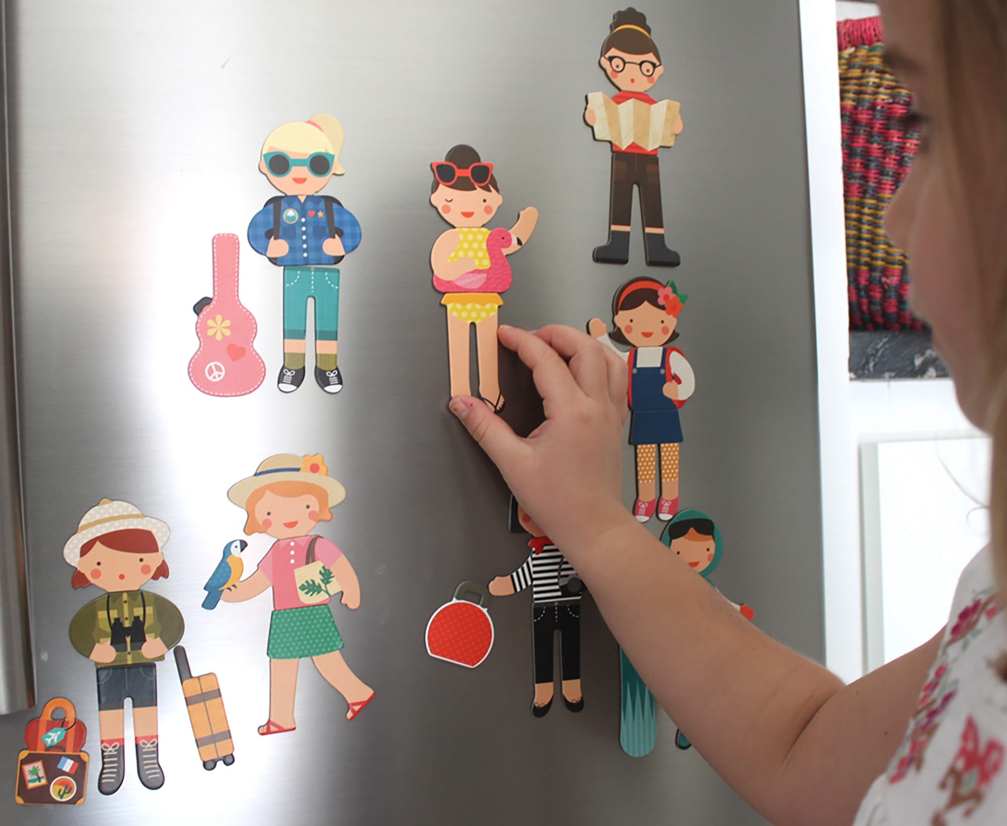 A person places a Petit Collage Little Travelers Magnetic Play Set doll on the fridge, surrounded by other stylish dolls and accessories from the set.