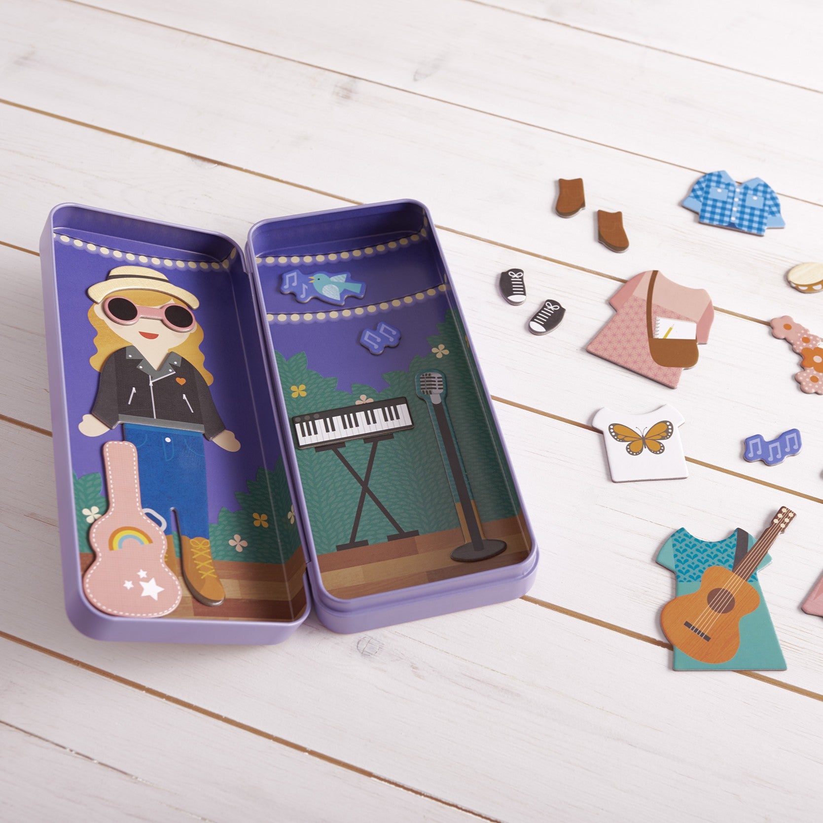 The Petit Collage Music Maker Shine Bright Magnetic Play Set includes a musician character, musical instruments, and mix-and-match clothing pieces, all arranged on a white wooden surface.