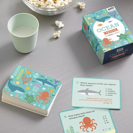 A set of Petit Collage Ocean Trivia cards is spread out on a table beside a cup of milk, a bowl of popcorn, and the game’s box.