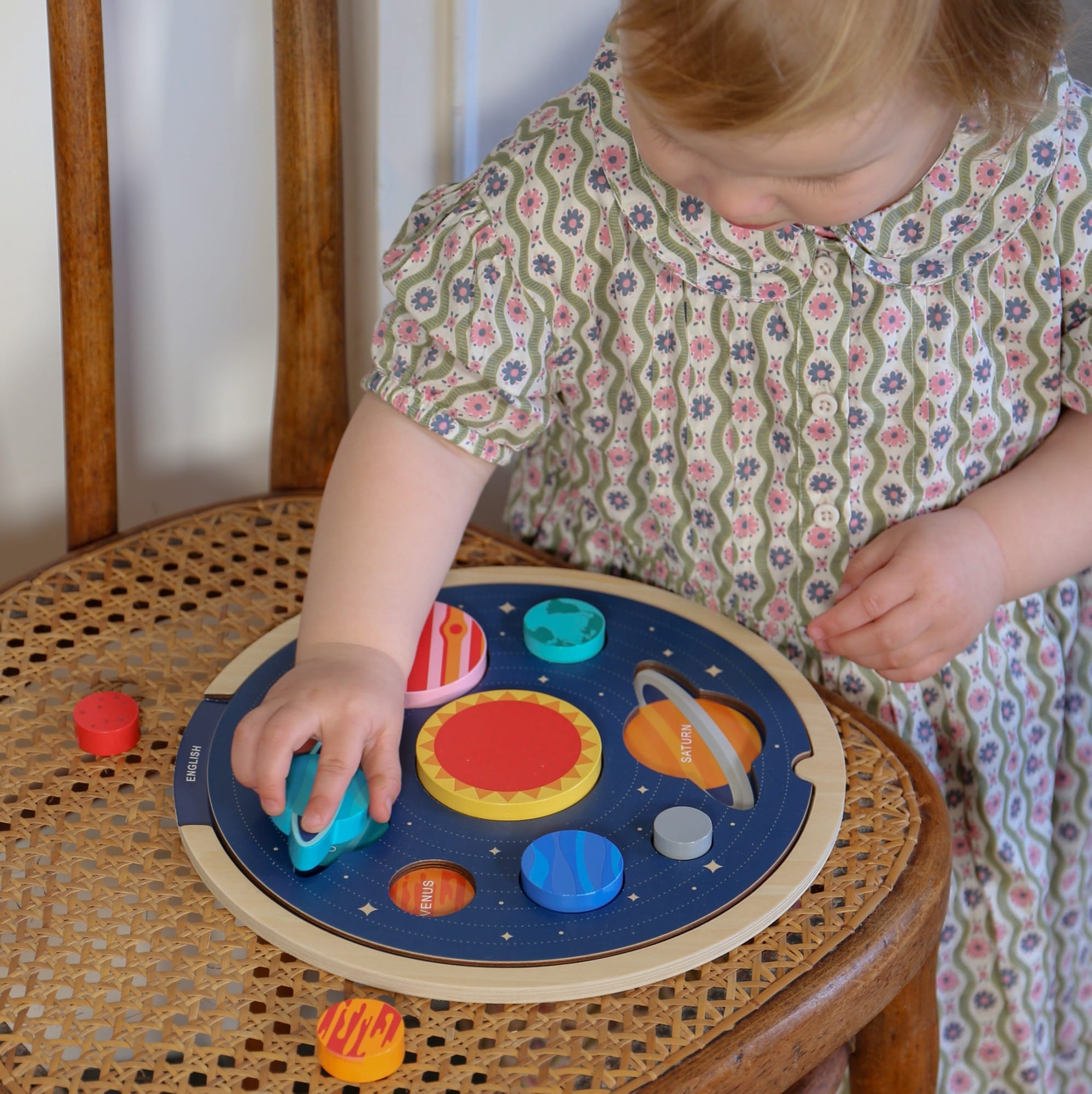A young child places wooden planet pieces onto the Petit Collage Multi-Language Solar System Wooden Tray Puzzle, which rests on a wicker chair.