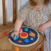 A young child places wooden planet pieces onto the Petit Collage Multi-Language Solar System Wooden Tray Puzzle, which rests on a wicker chair.
